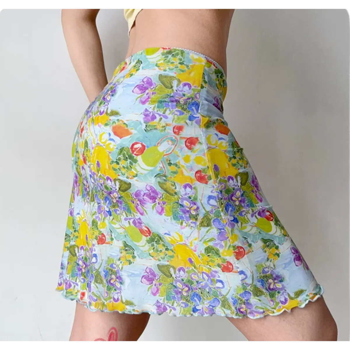 Side view of a light blue mesh mini skirt featuring a vibrant watercolor print of lemons, fruits, and purple flowers, with lettuce-edge hem and waist bow detail.