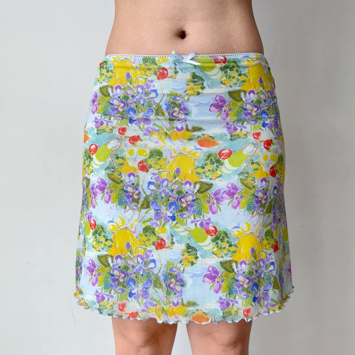 Close-up of a light blue mesh mini skirt, highlighting the watercolor fruit-and-floral print, waistband bow, and lettuce-edge hem.