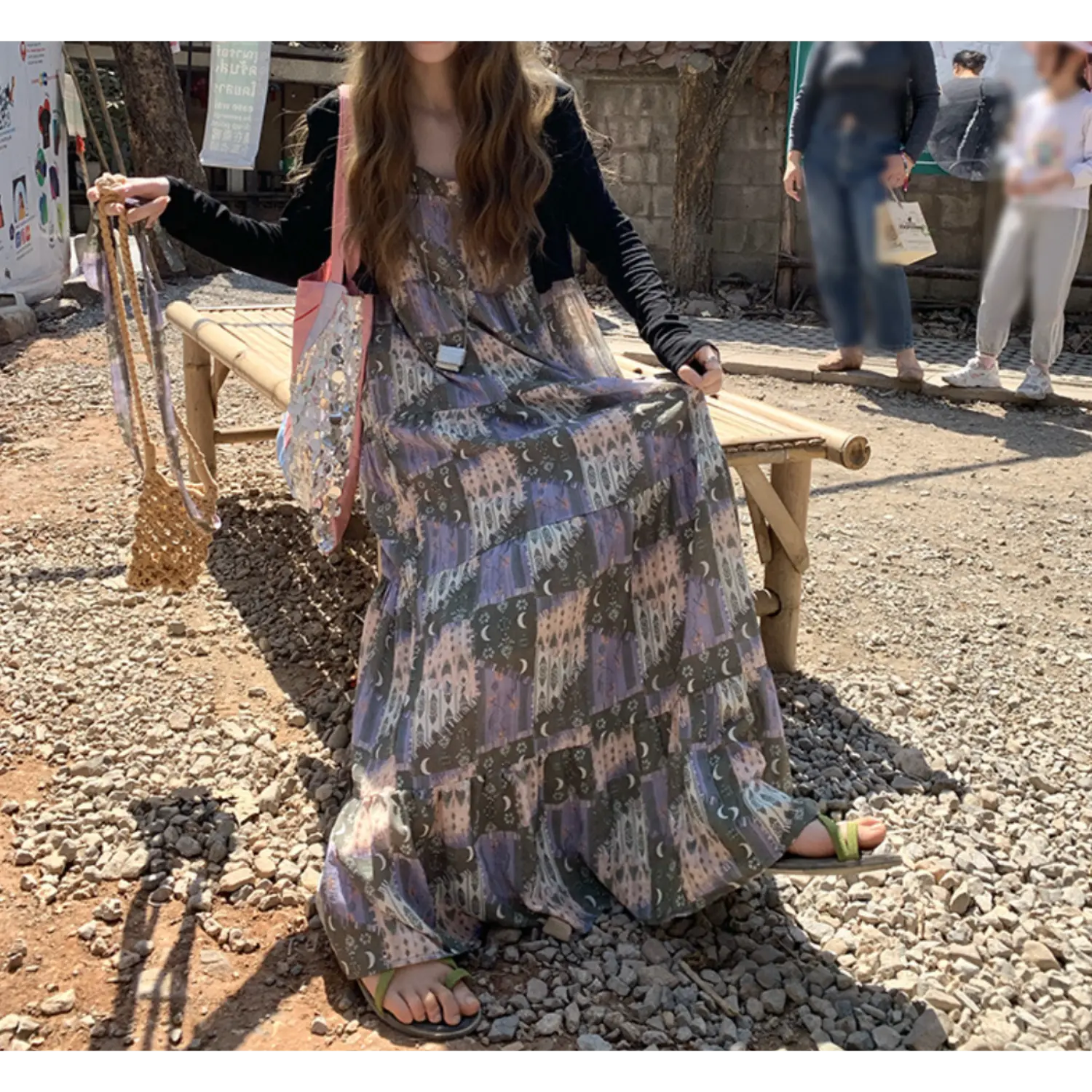 Outdoor gravel roadside sitting shot of loose tiered printed cami maxi dress layered with black long sleeve cardigan, vintage muted tone castle & moon print, ultra flowy multi-layer ruffle skirt, thin spaghetti straps, pink sequin canvas tote bag, woven straw shoulder bag, green slide sandals, bamboo bench & tropical foliage scenery