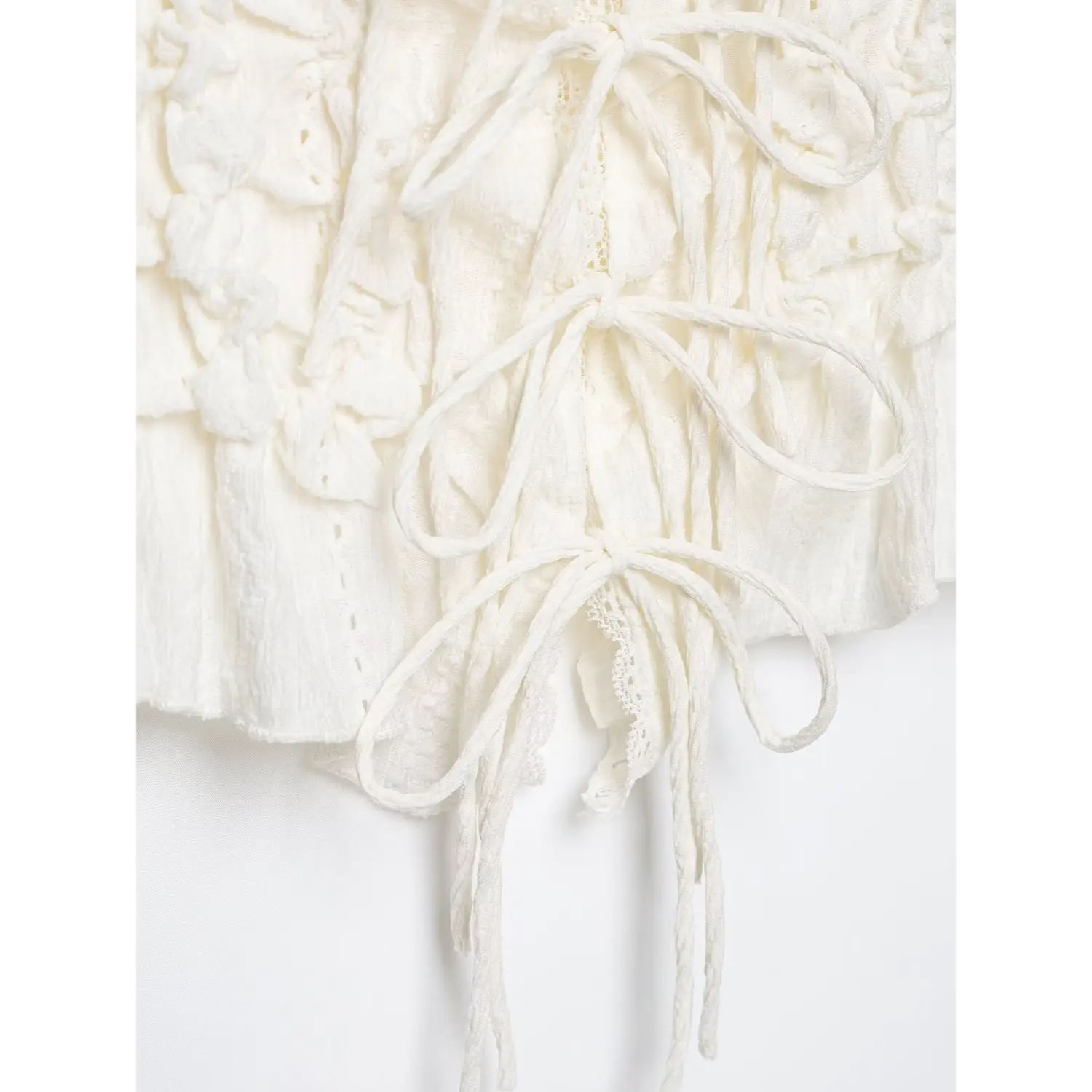 Close‑up bottom view of lace‑up ties and finished hem on white textured camisole top, loose hanging drawstring ties, white background