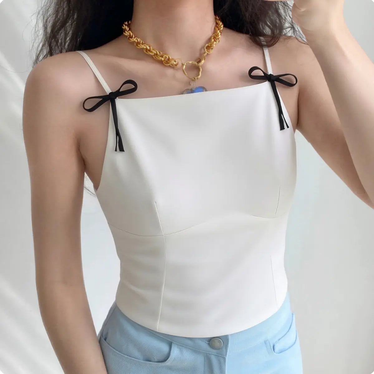 Lifestyle wearing shot of white camisole with black bow straps, paired with light blue denim jeans
