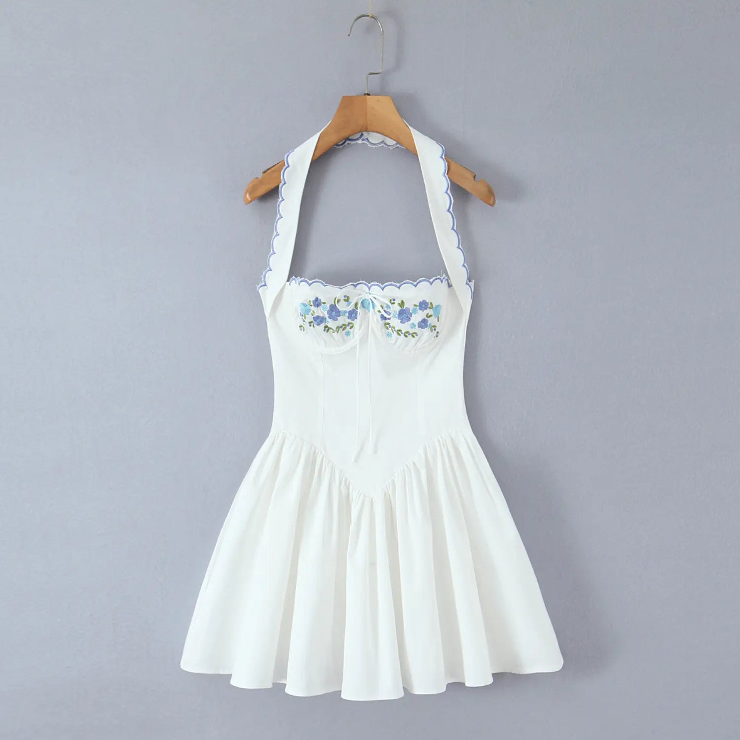 Hanging front shot, white cotton halter mini dress with blue floral embroidery, scalloped blue trim and tie bust detail