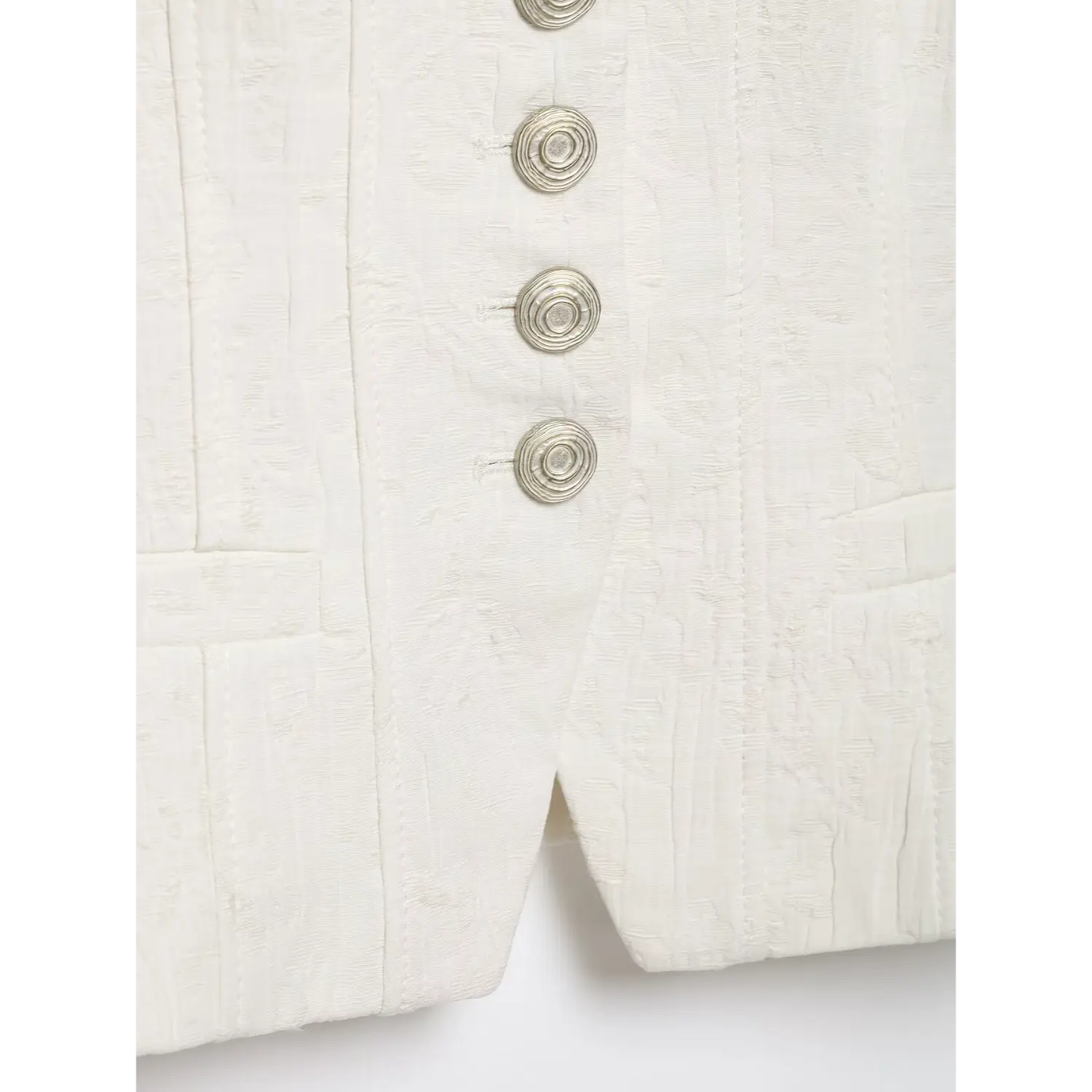 Close‑up detail of silver spiral button placket and front split hem on white textured cropped jacket, tailored seam lines, white background