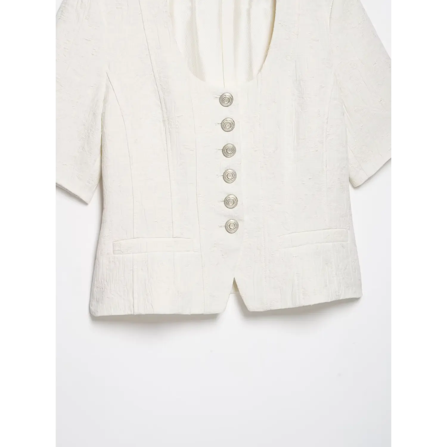 Partial full front shot of white textured cropped short sleeve jacket, u‑neck, silver spiral buttons, dual patch pockets, tailored seams, white background