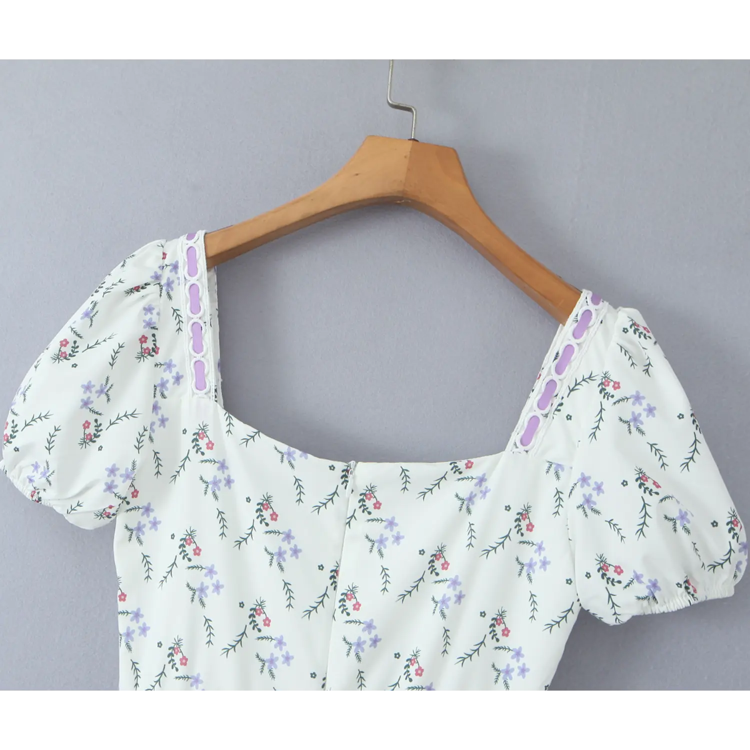 Back bodice close‑up of white ditsy floral mini dress, square neckline, puff short sleeves, purple eyelet trim, hanging on wooden hanger