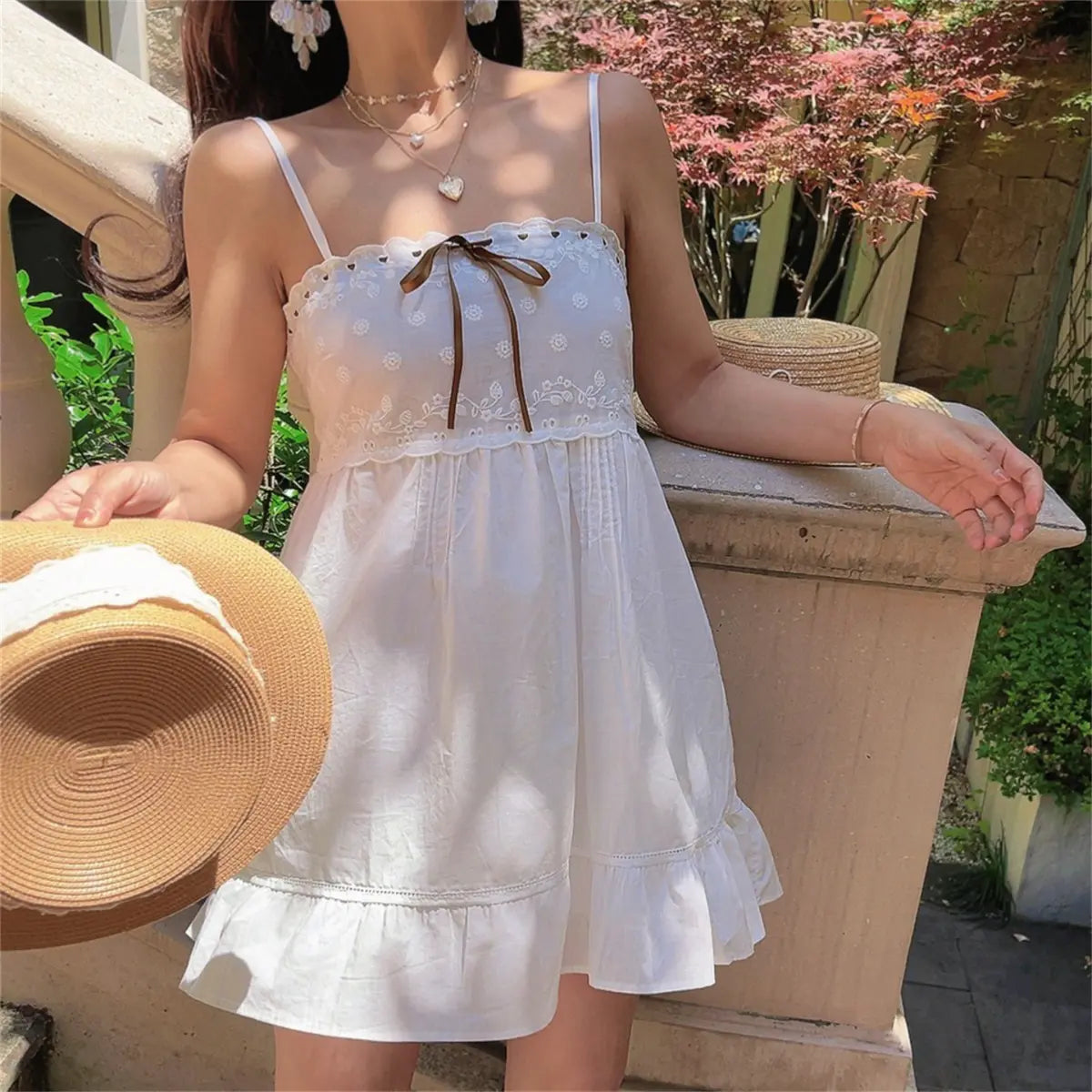 Outdoor shot of a model in a white embroidered cami dress, holding a straw hat and posing near a stone balustrade.