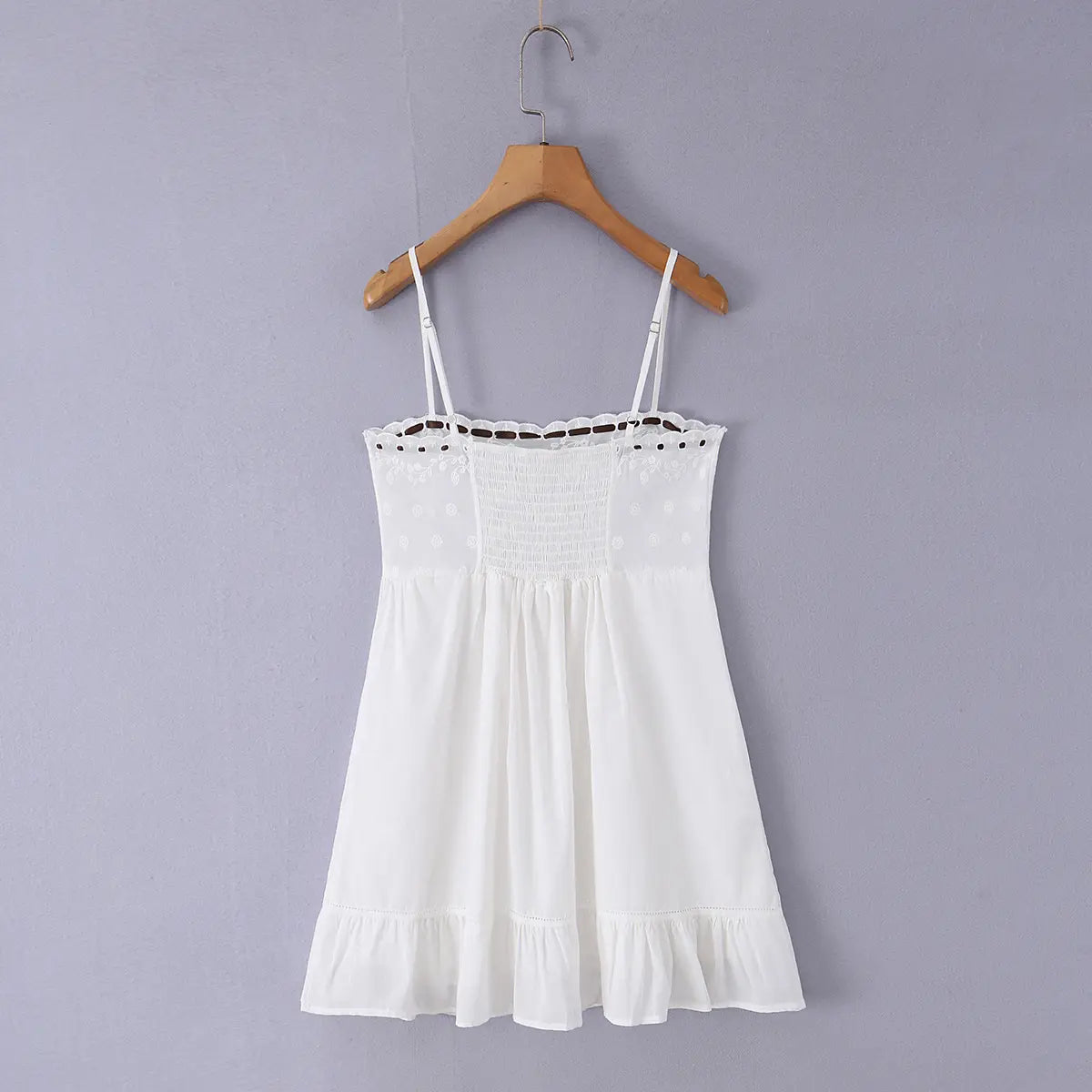 Full back view of a white embroidered cami mini dress, showing the smocked panel and ruffled hem, hanging on a wooden hanger.
