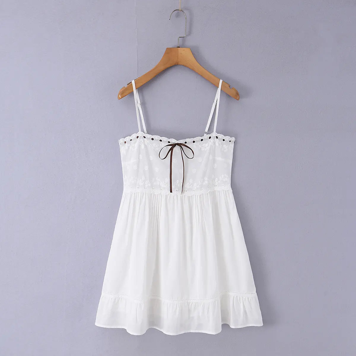 Full front view of a white embroidered cami mini dress with brown ribbon bow, scalloped neckline, and ruffled hem, hanging on a wooden hanger.