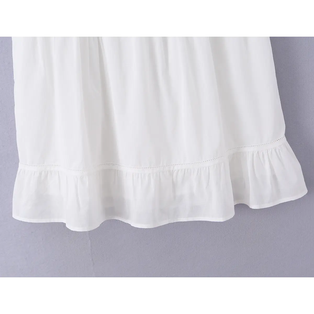 Close-up detail shot of the ruffled hem on a white embroidered cami mini dress, showing the eyelet stitching and gathered ruffle.