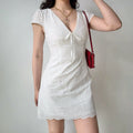 Full-body shot of a woman in a white eyelet embroidered mini dress, with one hand raised near her head