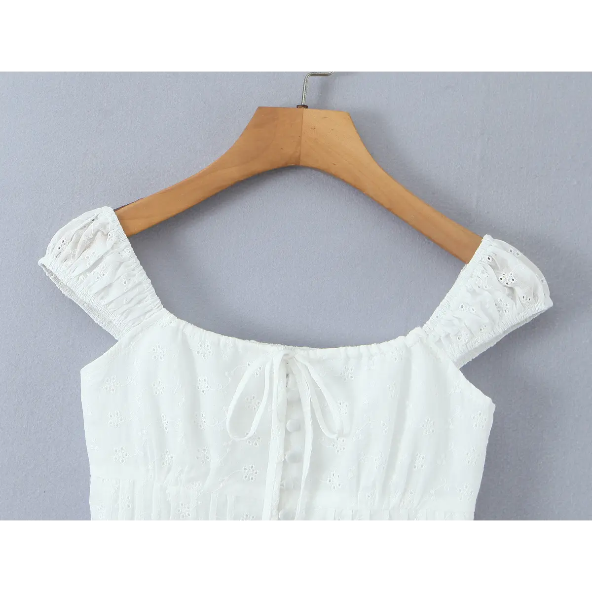 A white eyelet embroidered bustier-style top on a wooden hanger, featuring a square neckline, puff sleeves, and a front lace-up tie with button details.