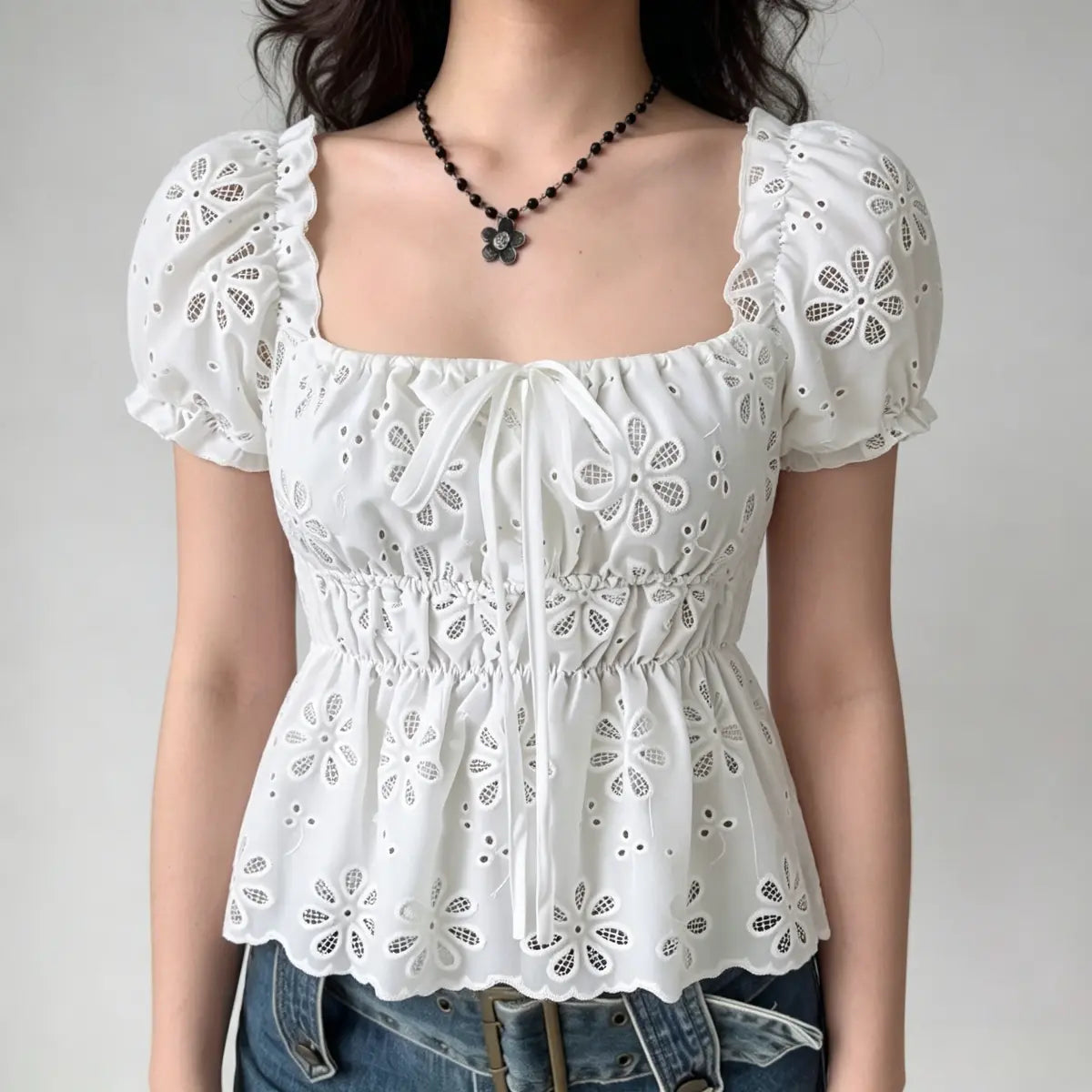 Model wearing a white eyelet-embroidered top with puffed sleeves, a square neckline, and a front tie detail, paired with jeans.