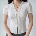 A woman wearing a white eyelet lace bustier shirt with a pointed collar, V-neck lace trim, puff sleeves, and two front tie closures, styled with gray bottoms and a pearl necklace.