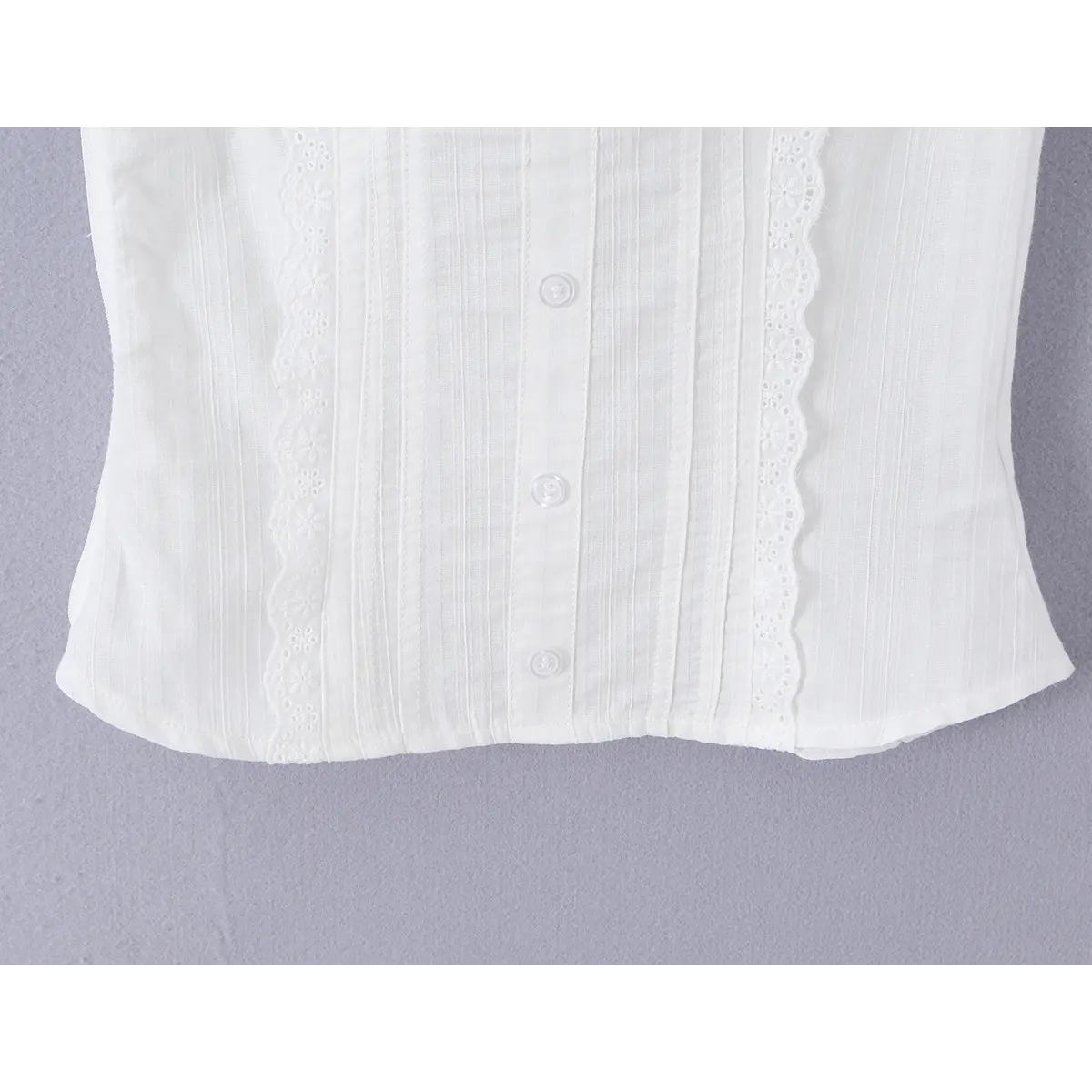 Close-up of the hem on a white ribbed bustier tank top, featuring vertical pintucks, eyelet lace trim, and front buttons.