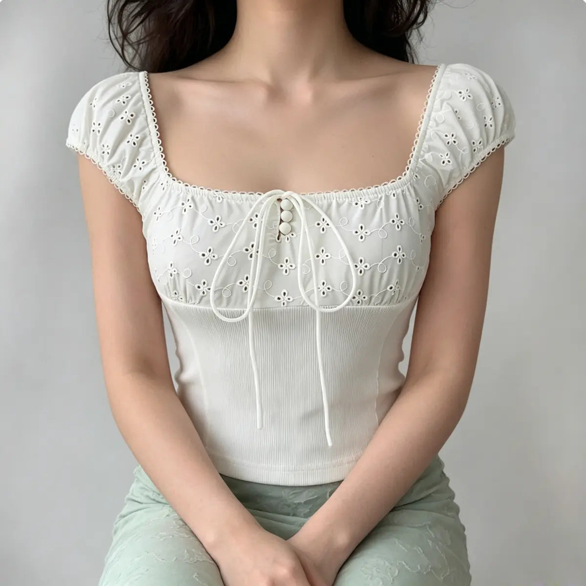 Model seated, wearing white eyelet embroidered top paired with green bottoms, showing the square neckline and fitted silhouette.