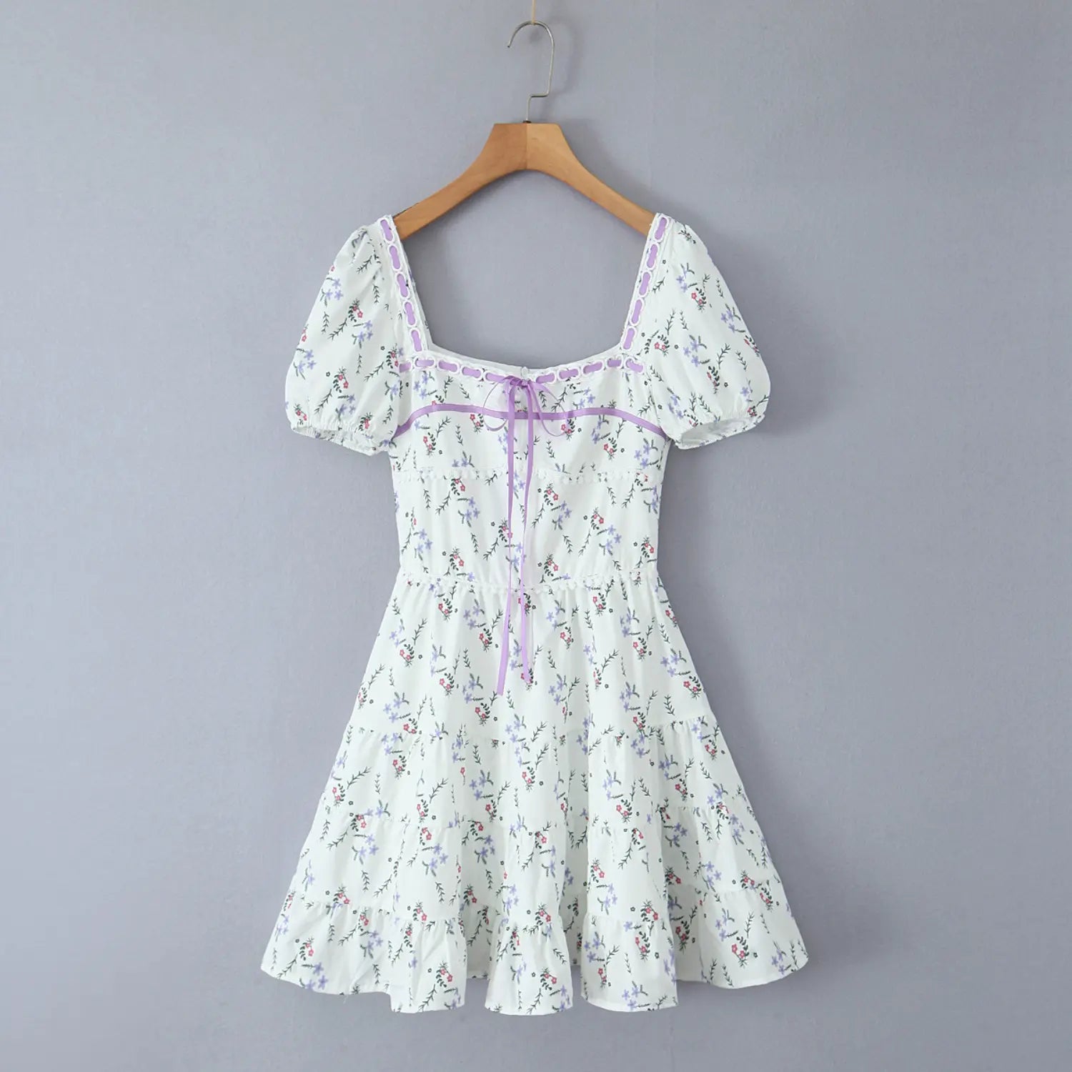 Full hanging front view of ditsy floral mini dress, purple ribbon lace‑up decor, tiered ruffle hem against grey wall