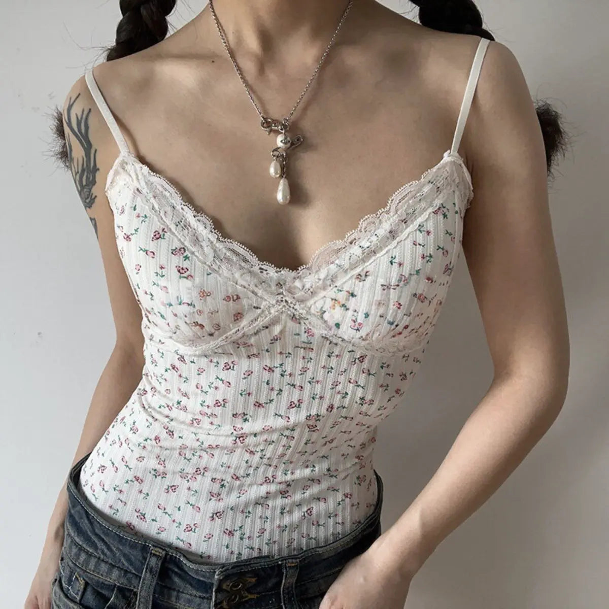 A woman wearing a white floral lace-trimmed cami top with denim jeans, accessorized with a pearl necklace.