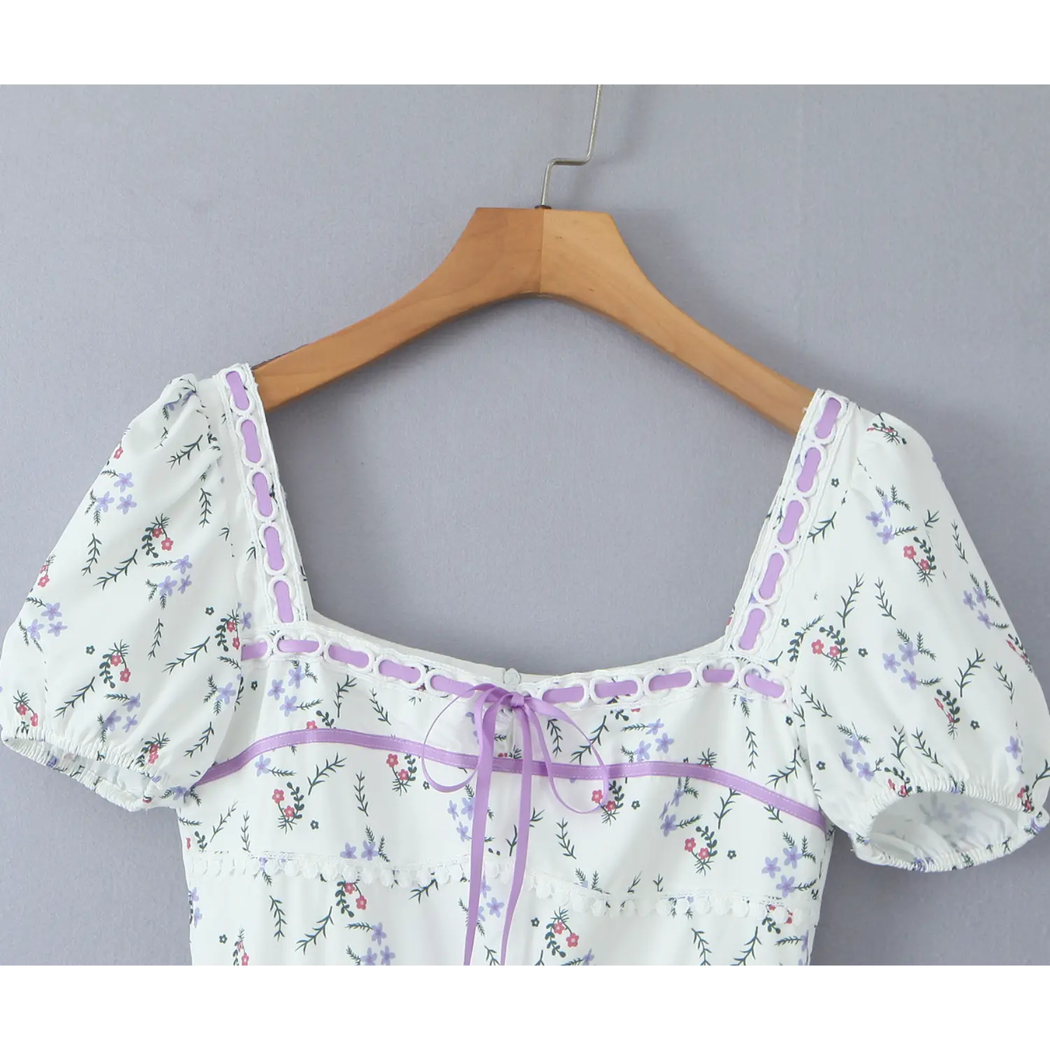 White ditsy floral mini dress hanging on wooden hanger, square neck, puff short sleeve, purple ribbon lace‑up detail on bodice