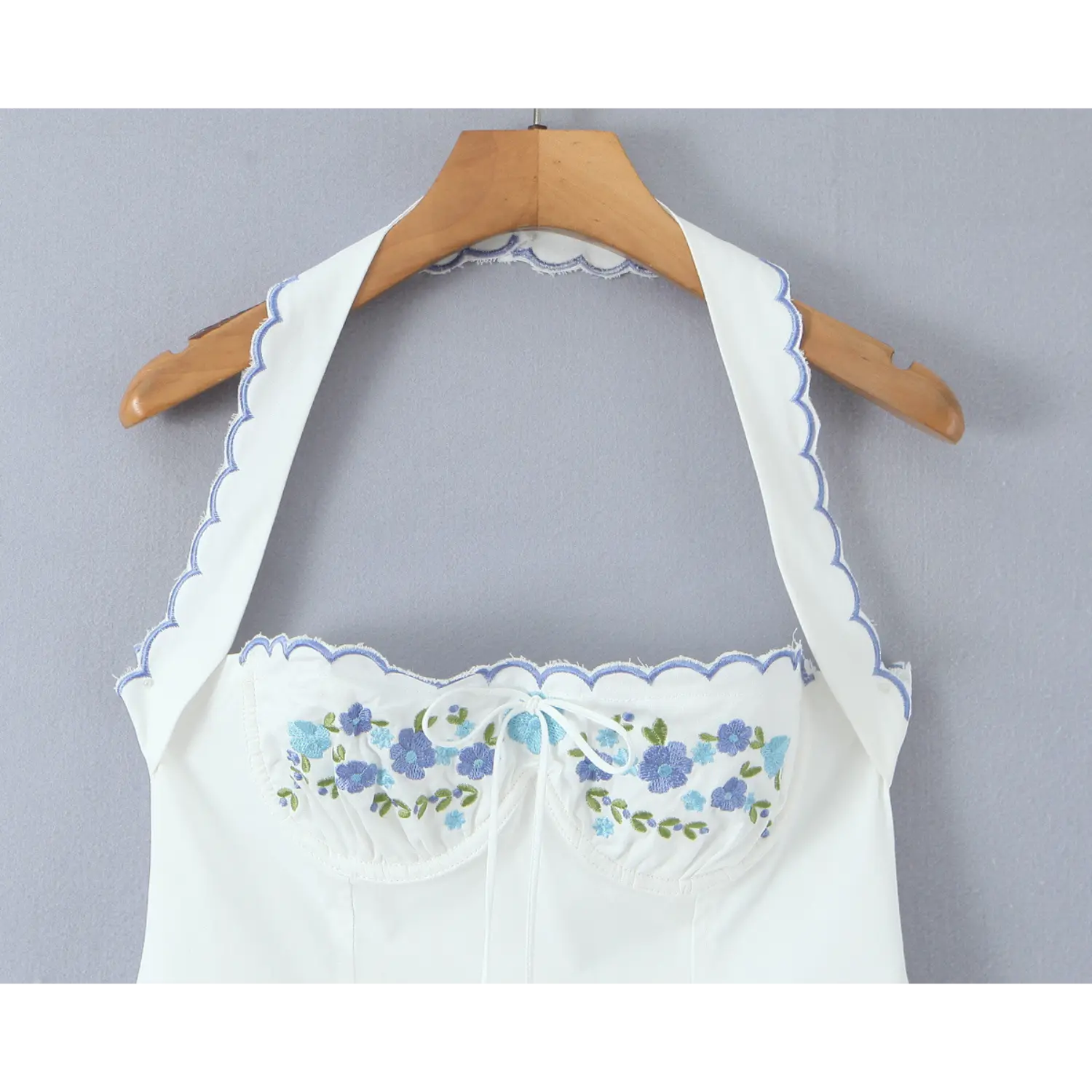 Closeup of bust section, white halter dress with blue floral embroidery, scalloped edge trim and front tie bow