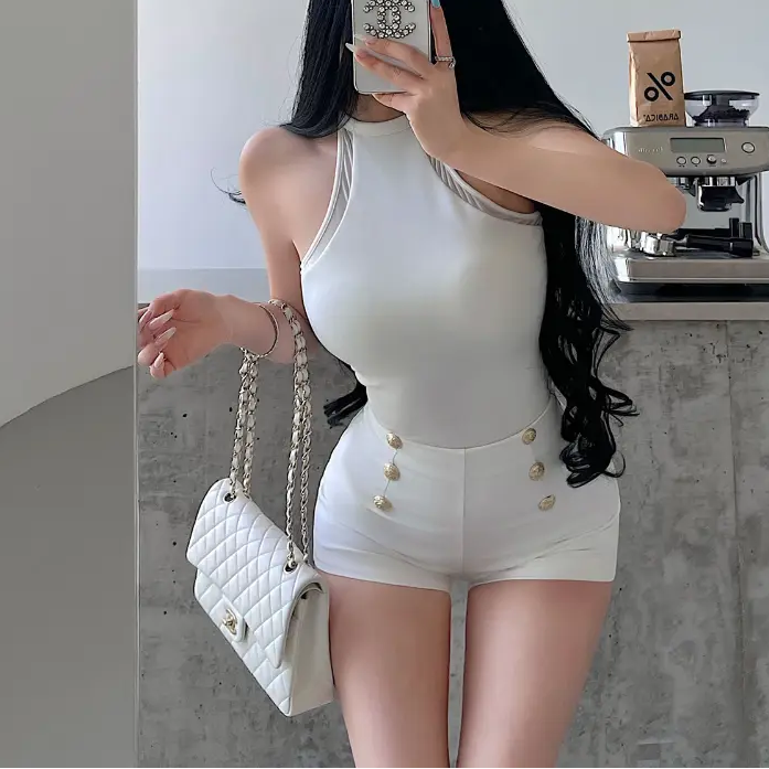 A woman wearing a white halter-neck romper with mesh panels and gold buttons, taking a mirror selfie.
