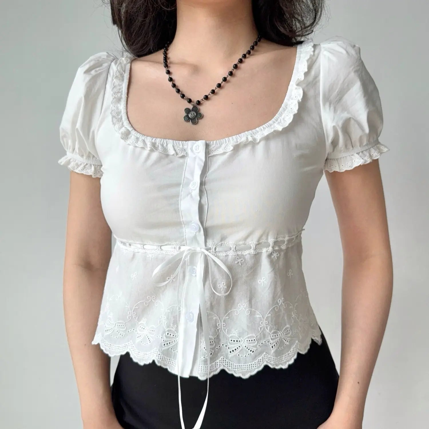 Close-up lifestyle shot of a woman wearing a white lace-trimmed puff sleeve top, showing the button placket and waist tie