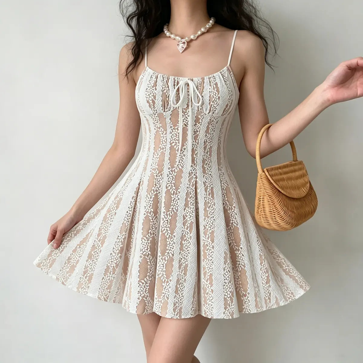 Model wearing a white lace spaghetti-strap mini dress with nude lining and center drawstring, splaying the skirt to show the flared silhouette, styled with a pearl necklace and wicker handbag.