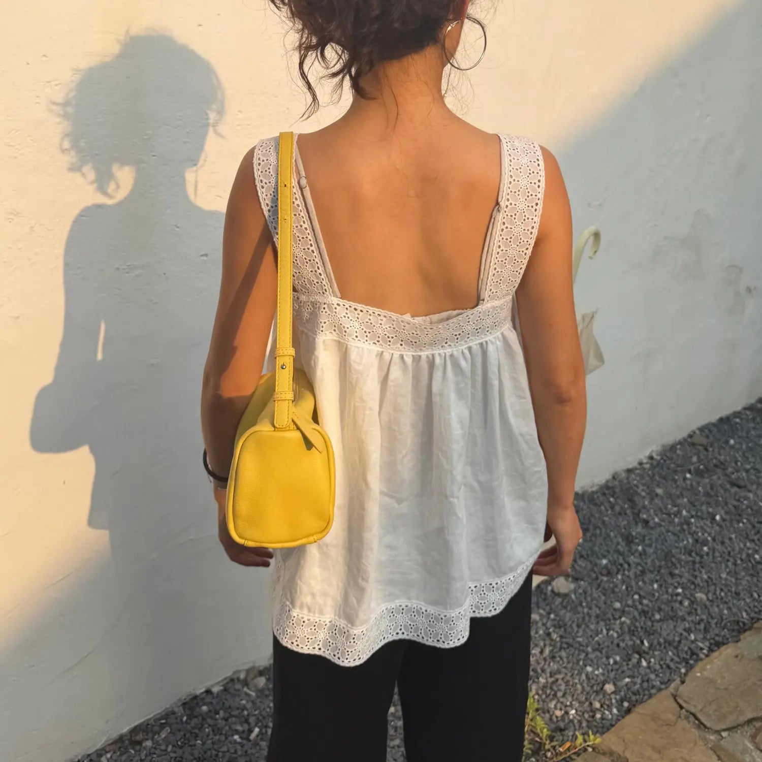 Back view of woman wearing white eyelet lace square neck camisole top, loose linen tank top, carrying yellow crossbody bag outdoors
