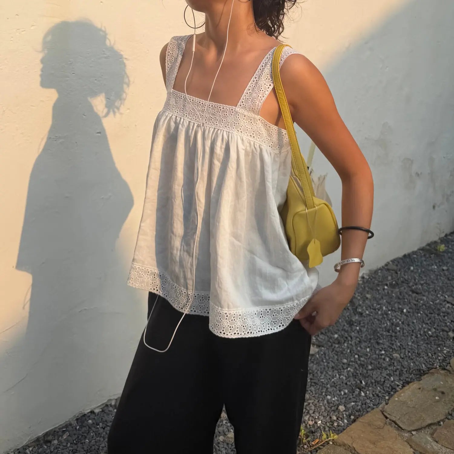 Side front outdoor shot of model in white eyelet lace sleeveless camisole, casual loose summer tank top matched with black pants