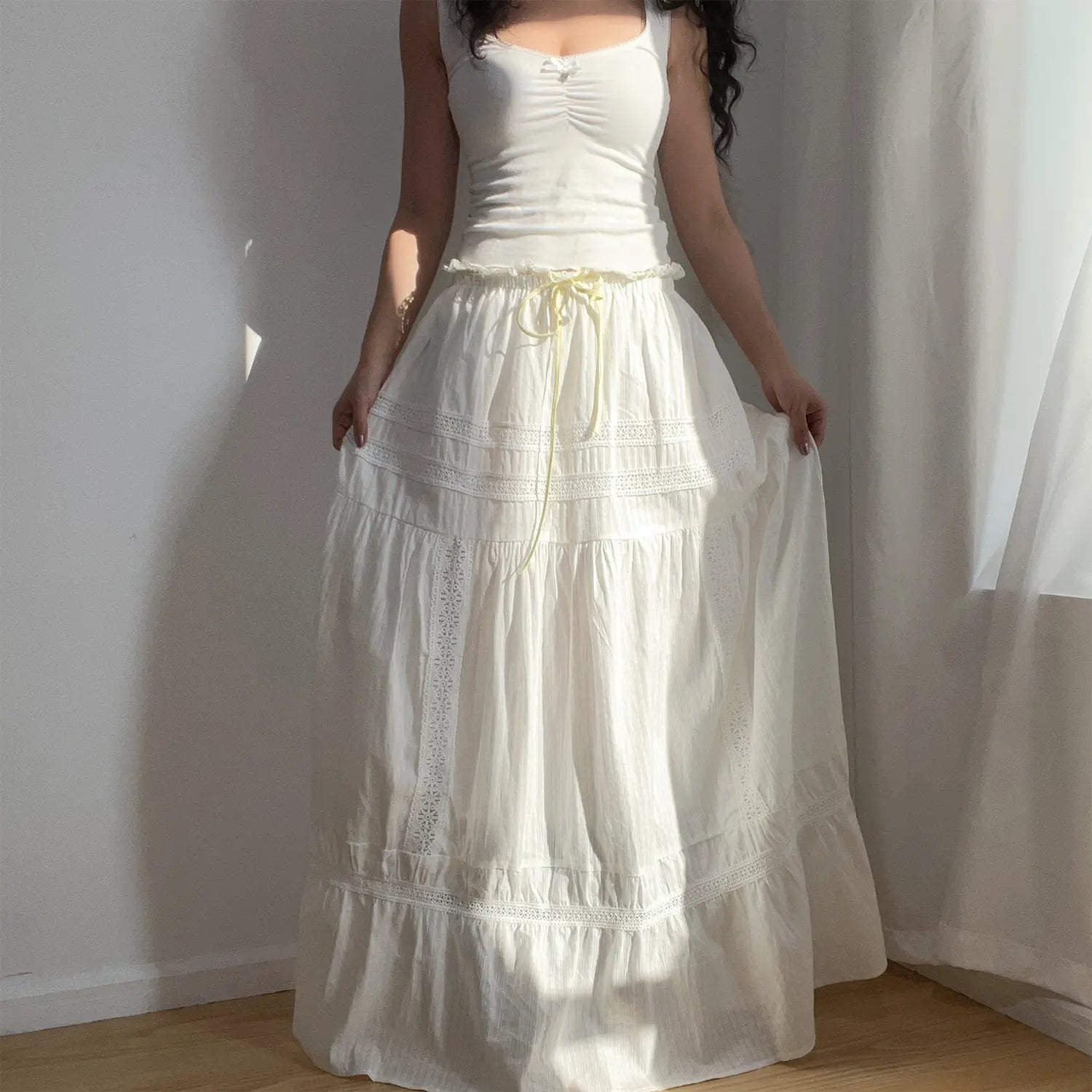 Full-body shot of a woman wearing a white lace tiered maxi skirt with a white tank top, standing by a sunlit wall.