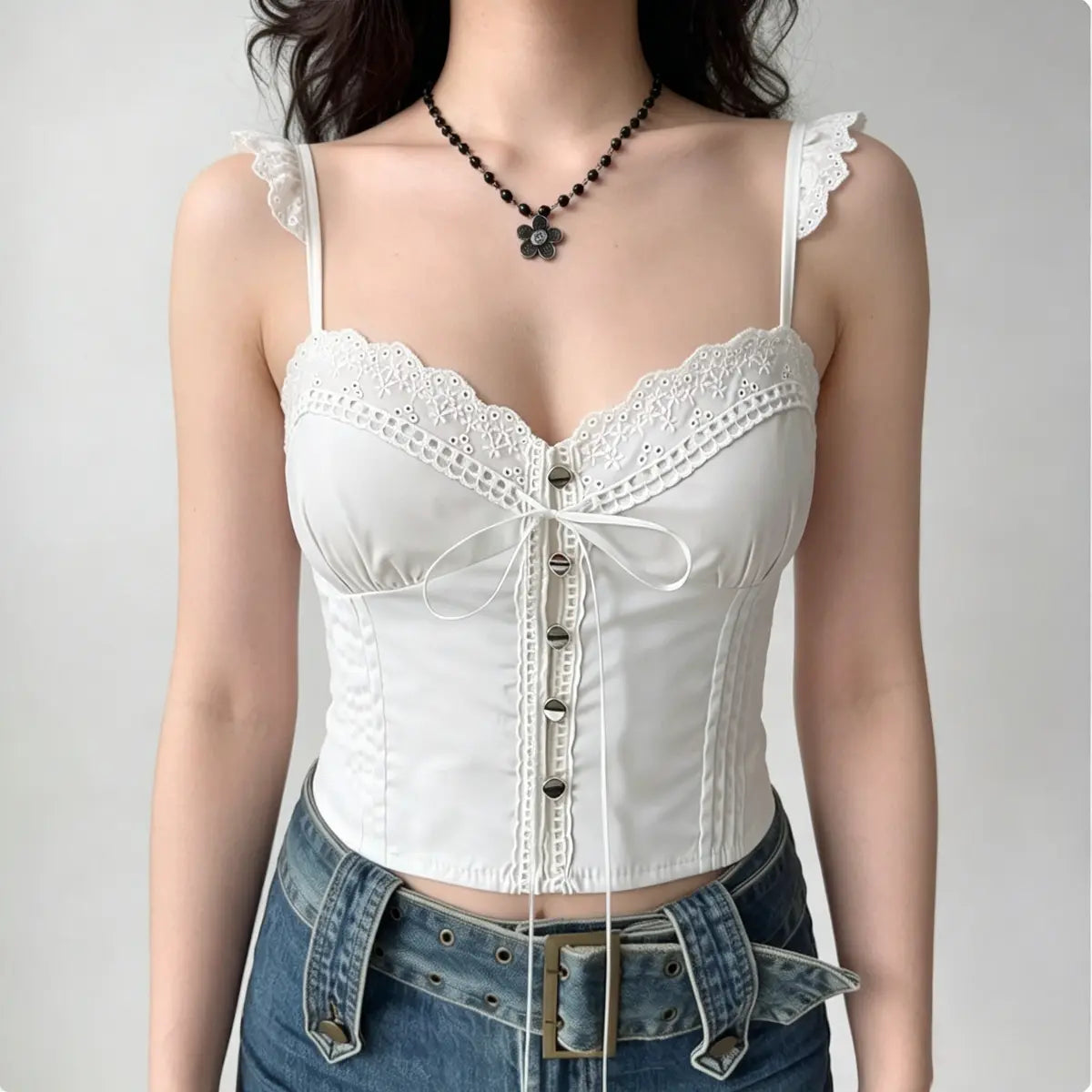 Model wearing a white lace-trimmed corset-style camisole top with a front bow, button details, and lace ruffled straps, paired with jeans.