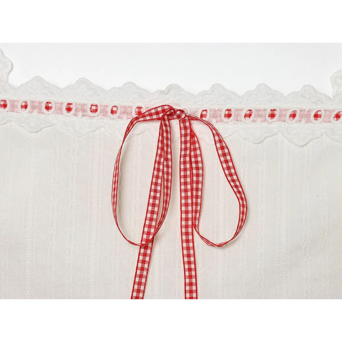 Close-up detail of the red gingham ribbon bow at the bust of a white lace-trimmed camisole, showing the lace and eyelet trim.