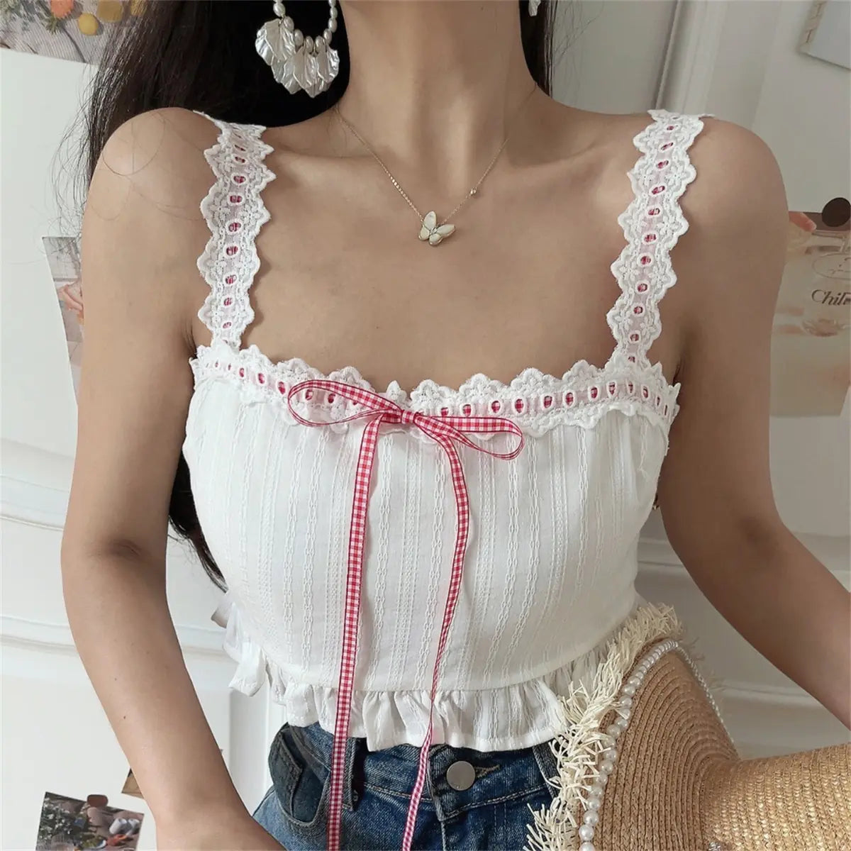 Front view of a woman wearing a white lace-trimmed camisole top with a red gingham ribbon bow at the bust, paired with jeans and holding a straw hat.