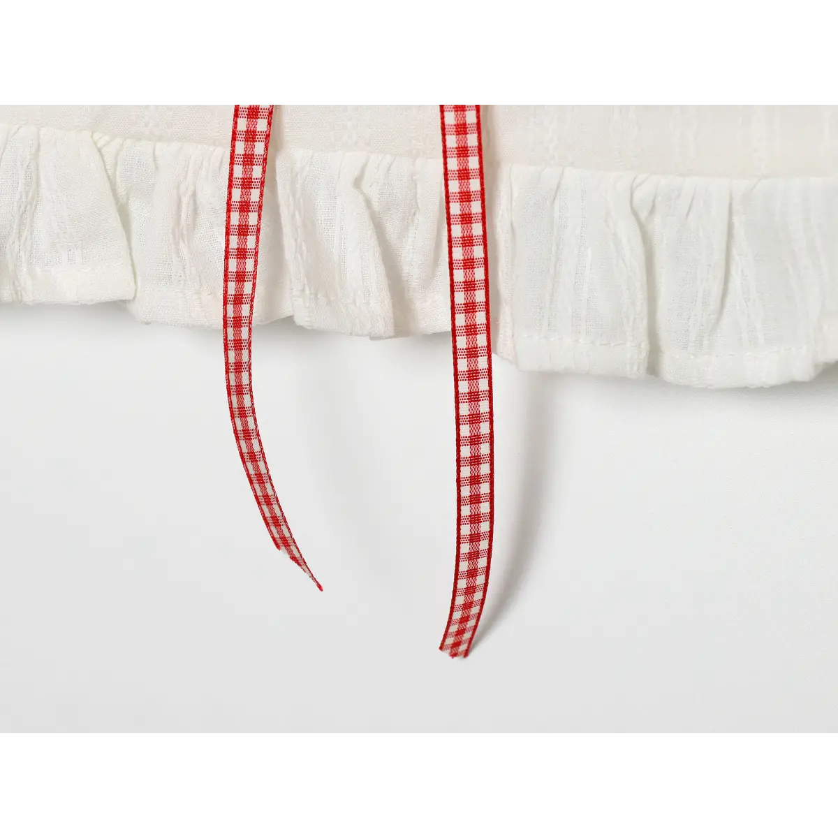 Close-up detail of the ruffled hem and red gingham ribbon ties on a white lace-trimmed camisole, isolated on a white background.