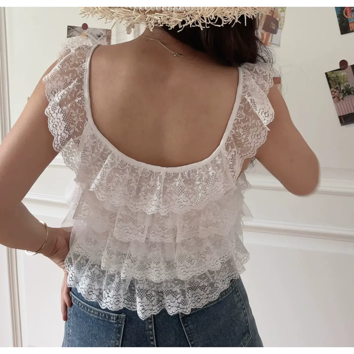 Back view of a woman wearing a white layered lace camisole with ruffled straps, paired with jeans and a straw hat.