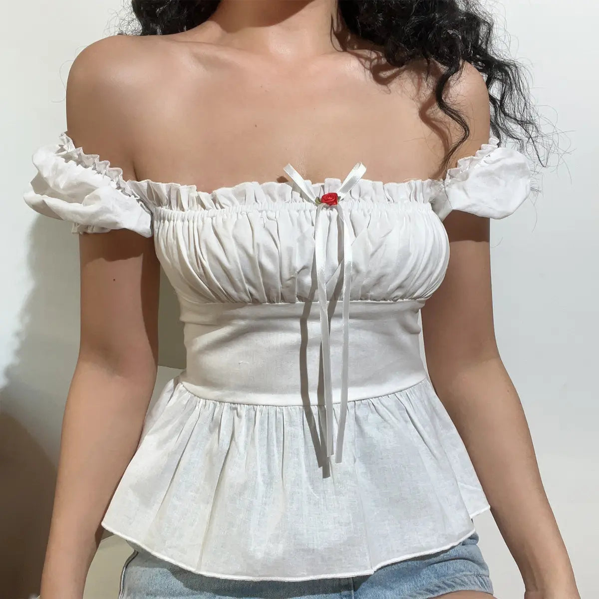 Model wearing a white off-shoulder peplum top with ruffled puff sleeves, featuring a gathered bust, ribbon bow with rose detail, and corset-style lace-up back.