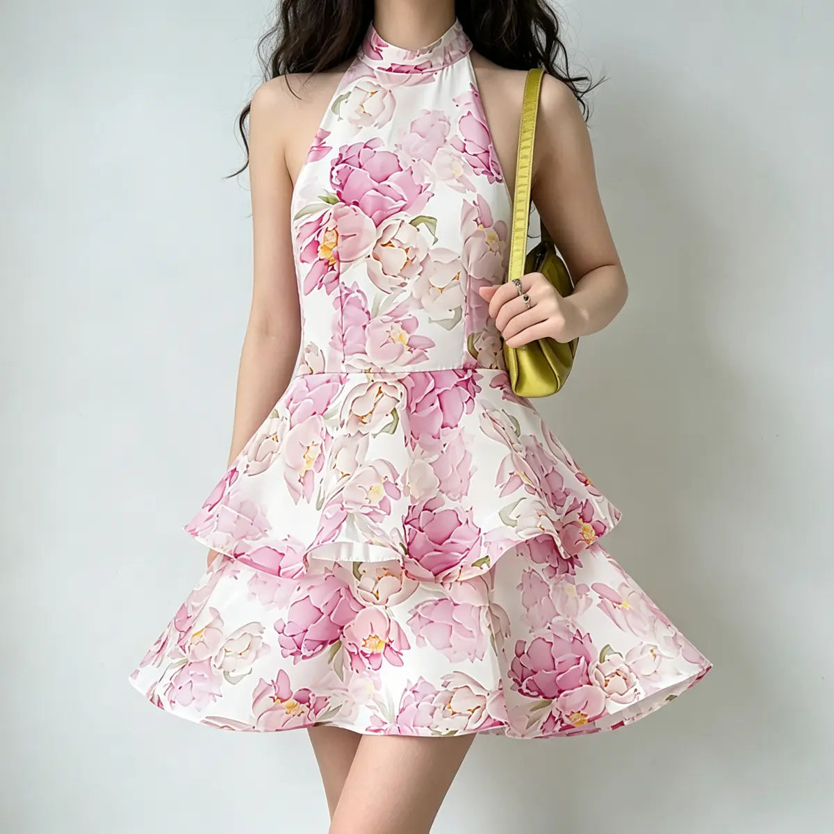 Model wearing a white halter-neck mini dress with pink peony floral print and tiered skirt, styled with a green satin handbag, casual pose.