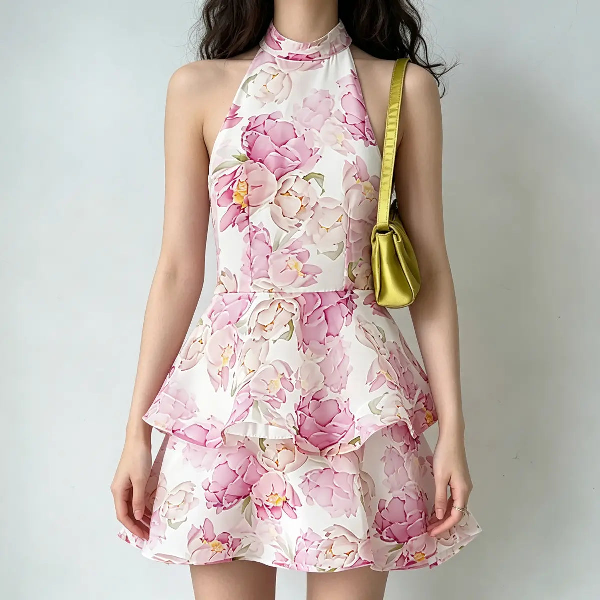 Model wearing a white halter-neck mini dress with pink peony floral print and tiered skirt, styled with a green satin handbag, front view.