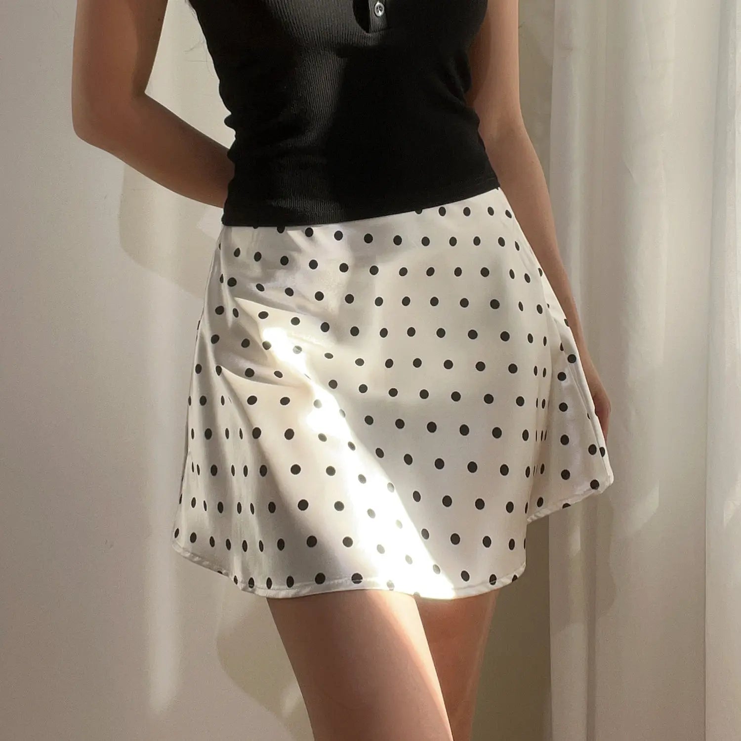 Female model wearing white black polka dot glossy satin high waist A-line mini skirt matched with black ribbed cropped polo top, indoor soft natural light shot