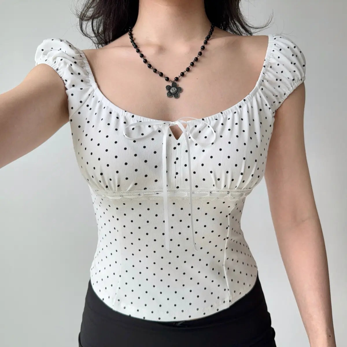 Woman wearing a form-fitting white polka dot top with puff sleeves, square neckline, and white ribbon tie detail at the bust.