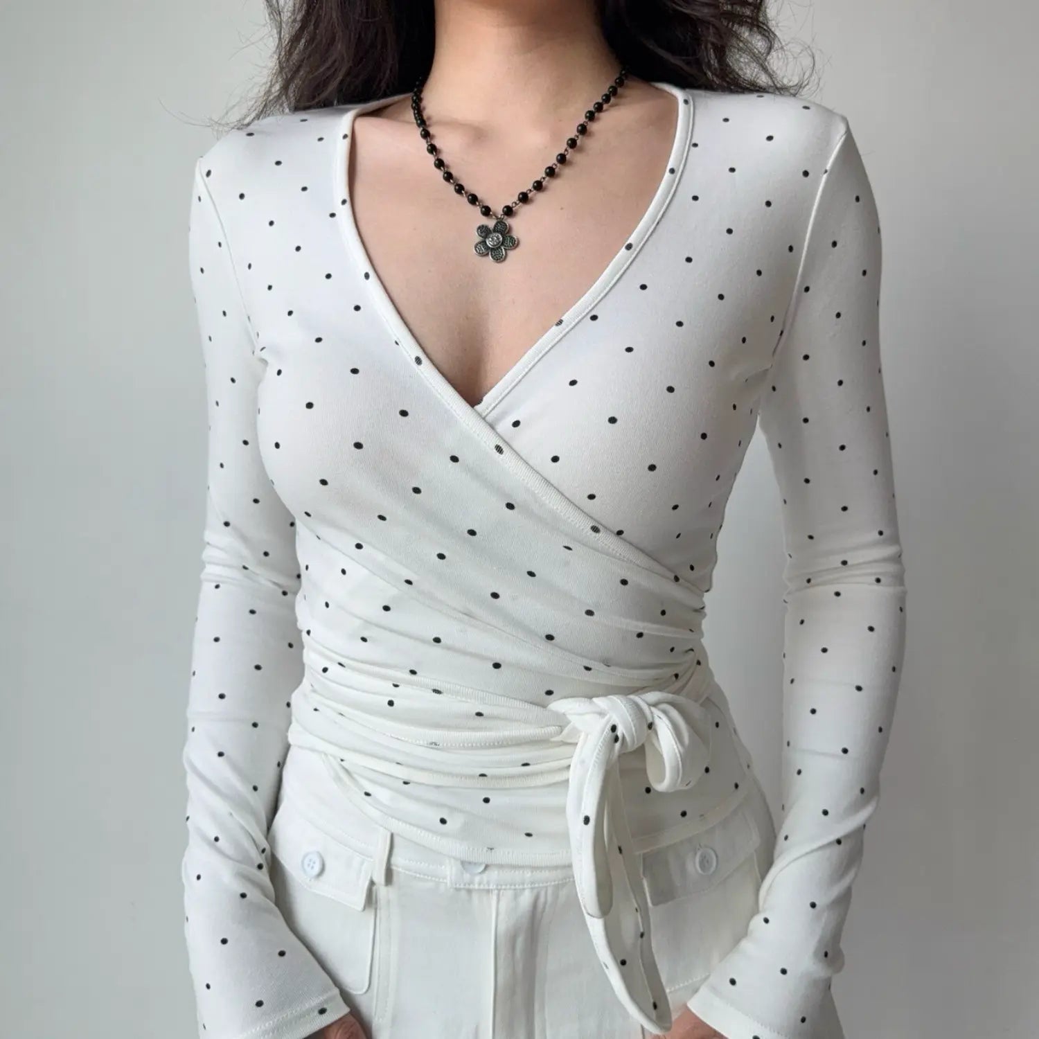 Front torso shot white polka dot wrap v‑neck long sleeve top with side waist tie knot fitted silhouette