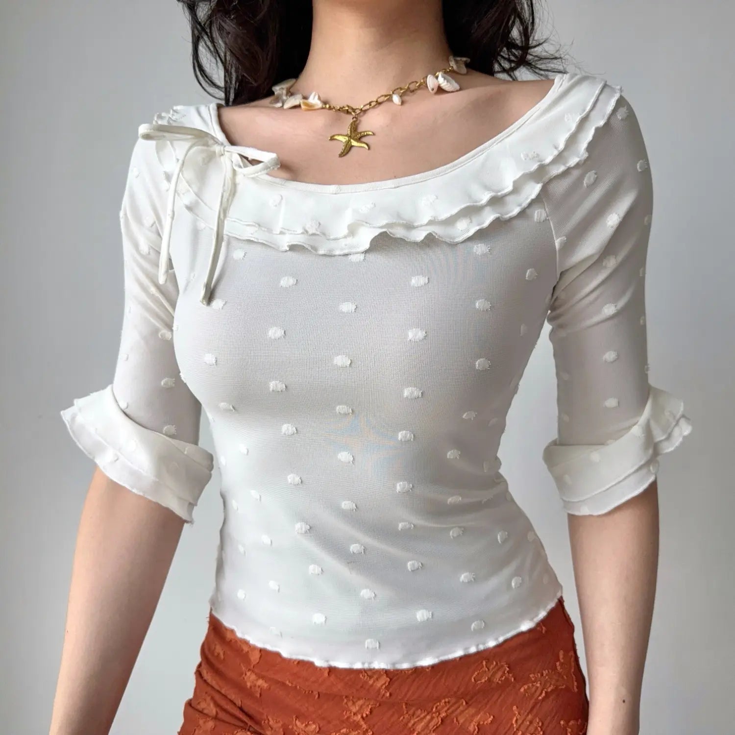 Front lifestyle shot on model of lightweight mesh fitted top, allover textured dots, layered neck frill, side bow decoration