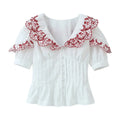 Front view of a white blouse with red embroidered ruffled bib collar, V-neck, button placket, pleated corset-style bodice, and puffed short sleeves.