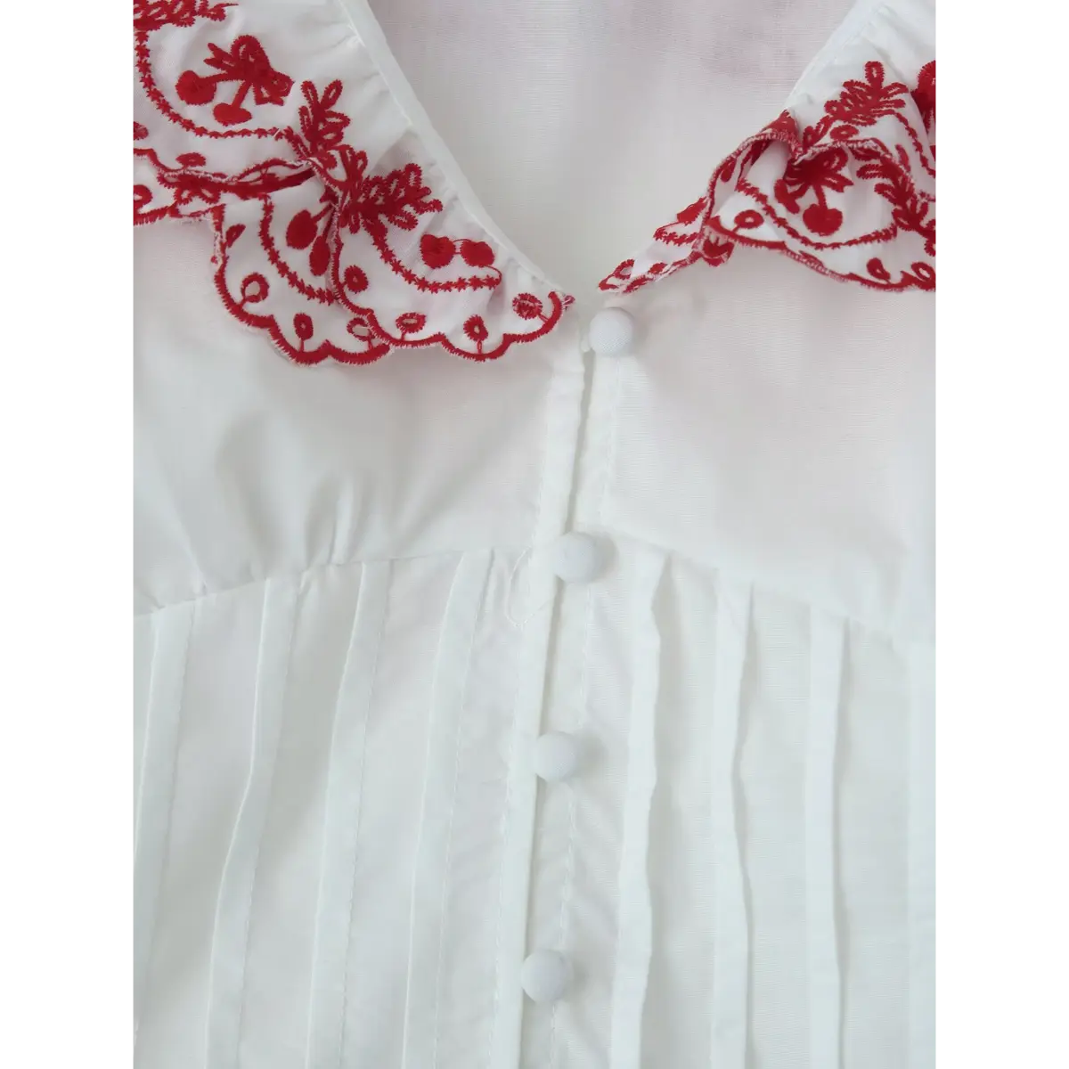 Close-up of the blouse's front placket, showing the round fabric-covered buttons, vertical pleats, and red cherry-embroidered ruffled collar.