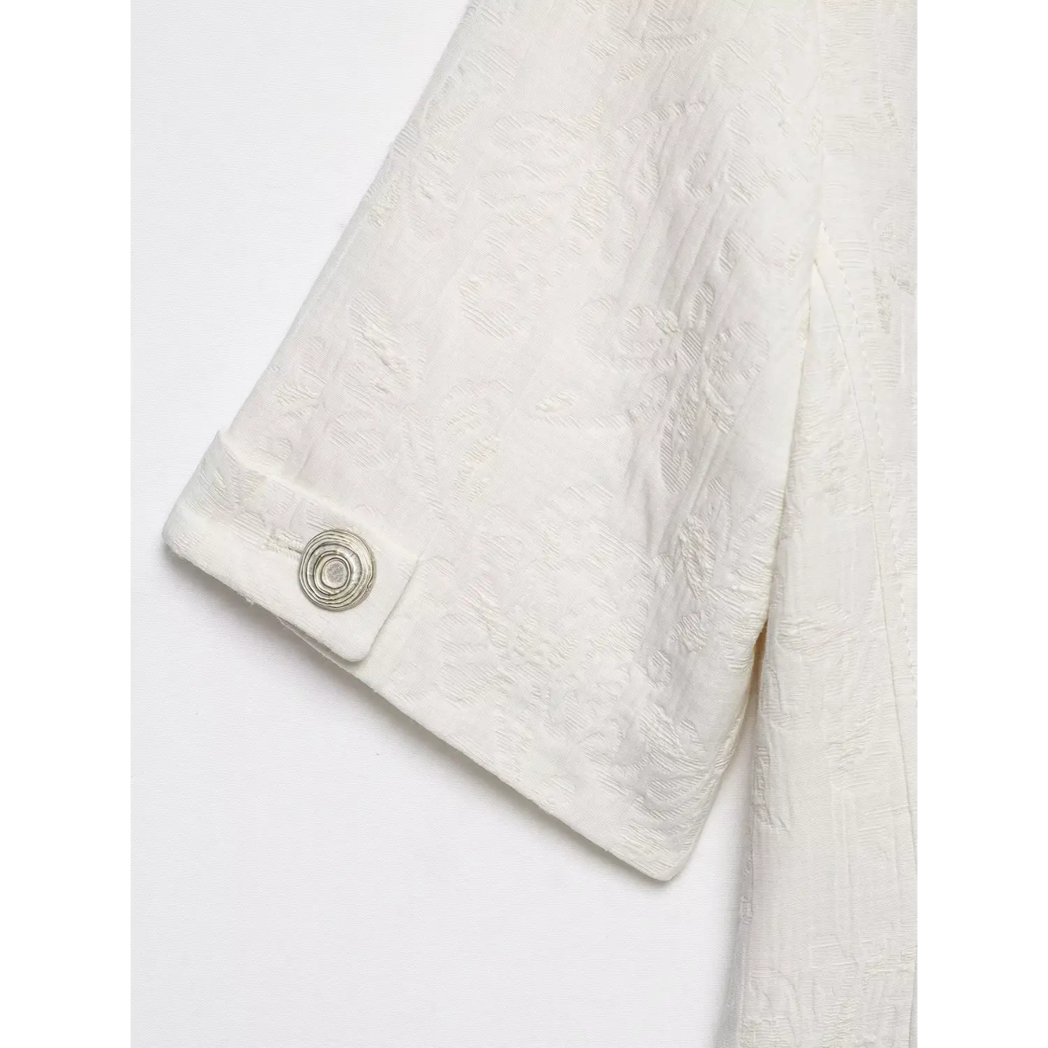 Close‑up of short sleeve cuff with decorative silver spiral button on white textured cropped jacket, embossed fabric texture, white background