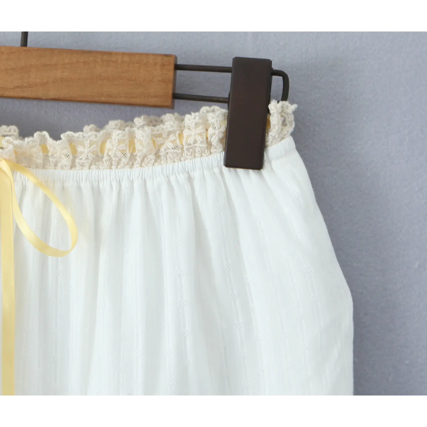 Close-up of the elasticated waistband of a white tiered maxi skirt, featuring a ruffled lace trim and a wooden hanger
