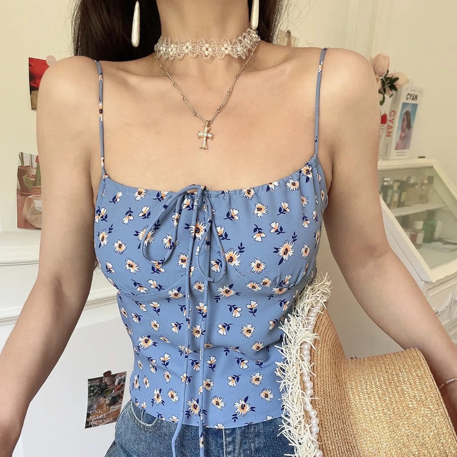 Women haze blue tiny daisy cami crop top, elastic square neck front drawstring tie, indoor half body model shot holding pearl frayed straw bag