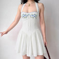 Model wearing white halter mini dress, blue floral bust embroidery, scalloped trim, fitted waist and flowy flared skirt