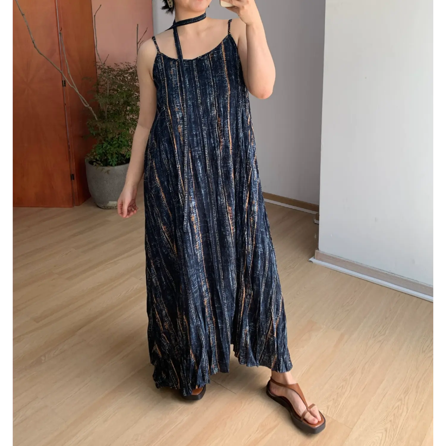 Three-quarter indoor mirror shot of loose A-line crinkle cami maxi dress, faded vertical bronze-blue stripe print, scoop neck thin spaghetti straps, long matching fabric choker tie, brown platform thong sandals, minimalist light wood floor & potted plant room decor