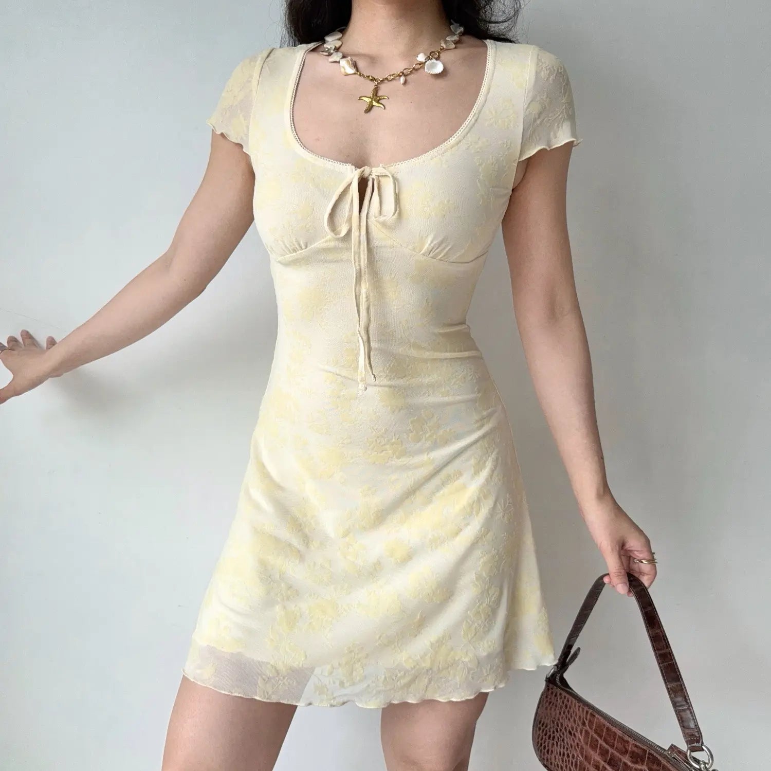Model front reach‑out pose yellow floral flocked mesh mini dress u‑neck tie bust detail short sleeve mesh dress