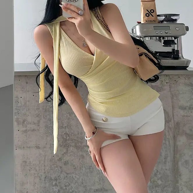 A woman wearing a yellow ribbed halter wrap top with a tie detail, styled with white shorts and a straw hat, taking a mirror selfie.
